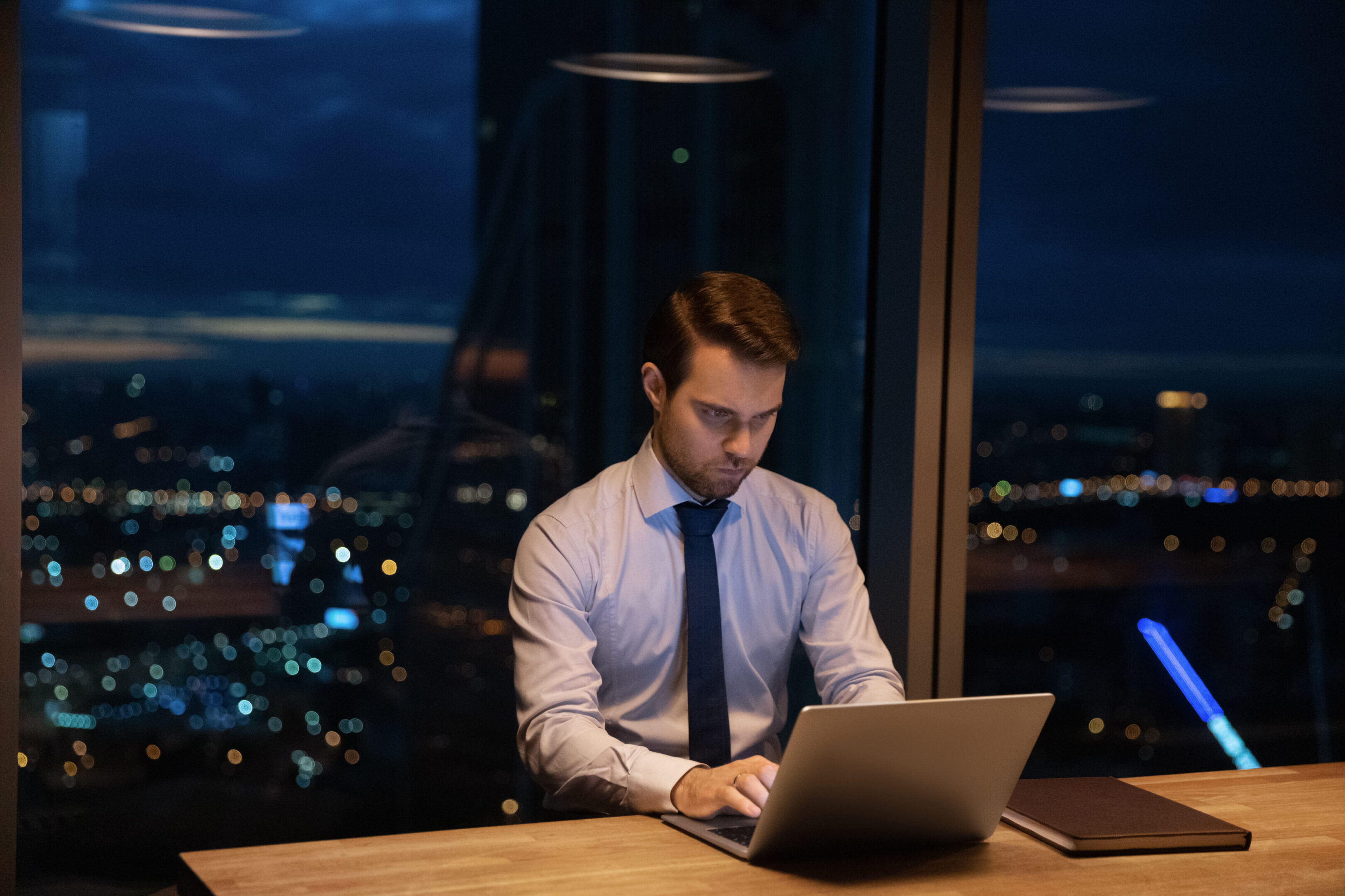 Confident ambitious corporate male office employee sit at workplace work until late use laptop, lead correspondence to client distantly, make urgent task. Workaholism, careerism, after-hours concept