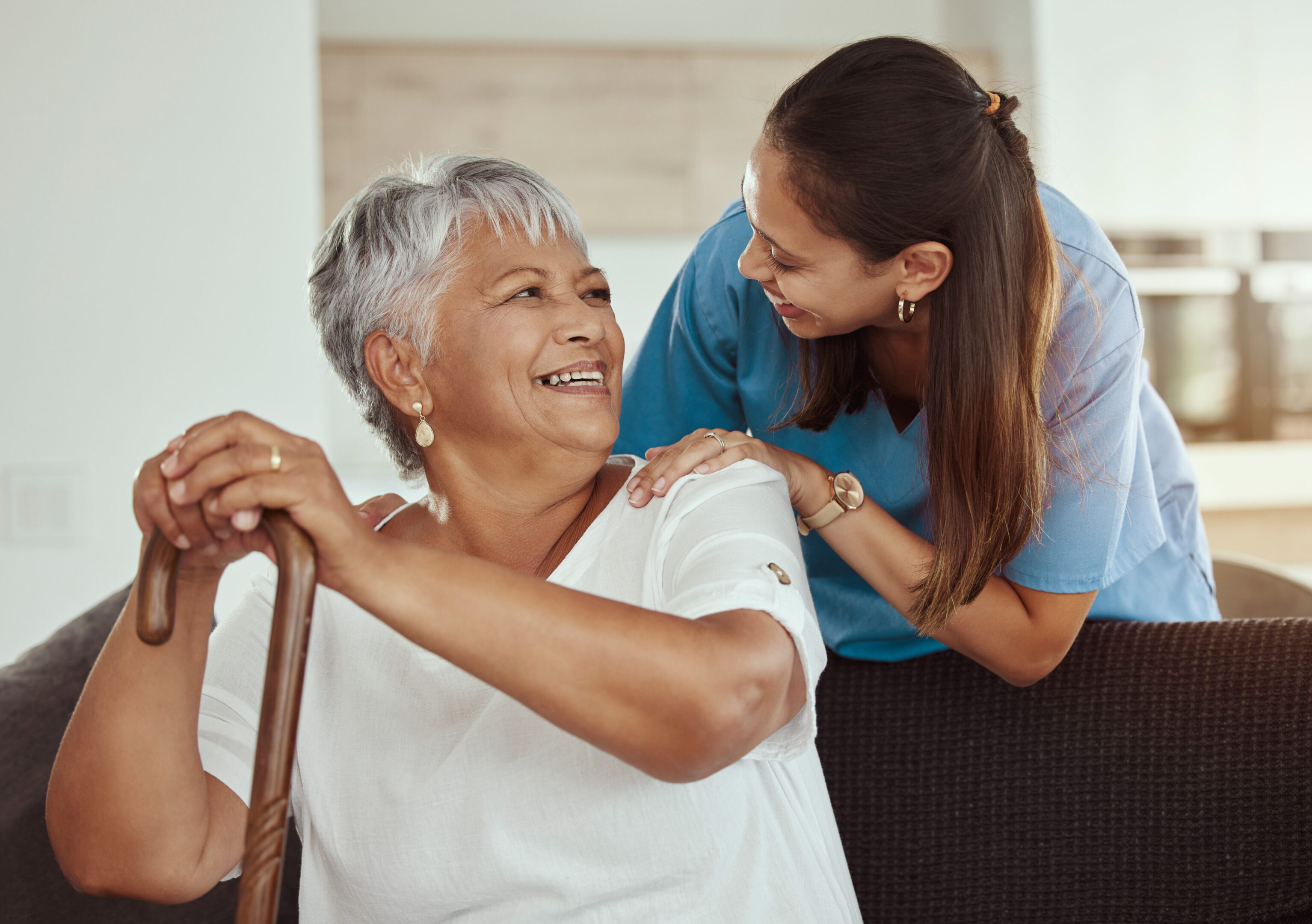 Happy, relax and senior woman with caregiver smile while sitting on a living room sofa in a nursing home. Support, help and professional nurse or healthcare worker helping elderly lady or patient